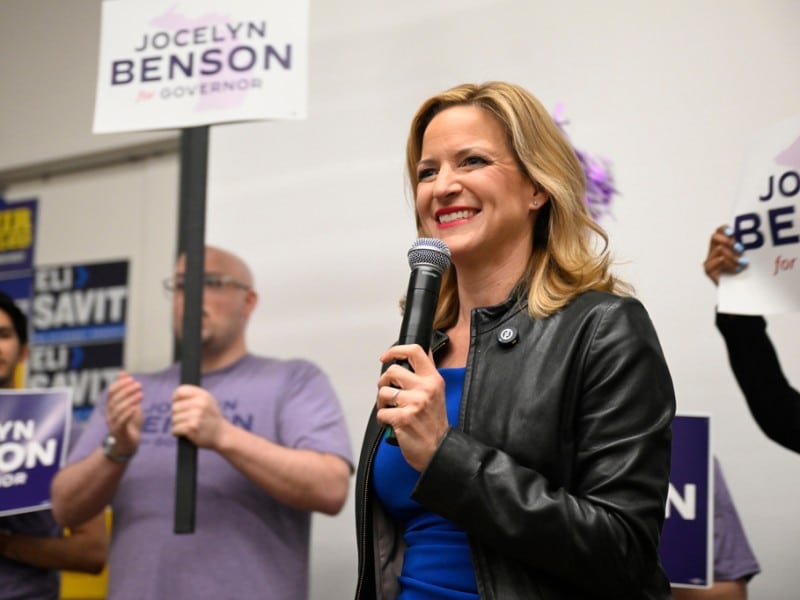 Jocelyn Benson smiles while speaking into a microphone, surrounded by supporters holding "Jocelyn Benson for Governor" signs.
