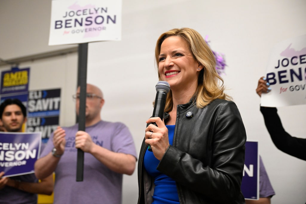 Jocelyn Benson smiles while speaking into a microphone, surrounded by supporters holding "Jocelyn Benson for Governor" signs.