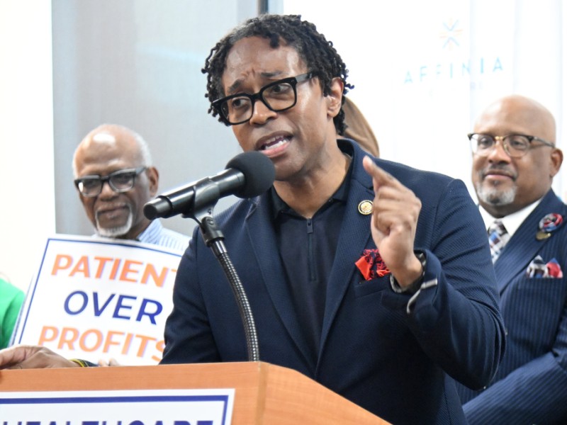 U.S. Rep. Wesley Bell speaks into a microphone at a podium next to a "Patients Over Profits" sign.