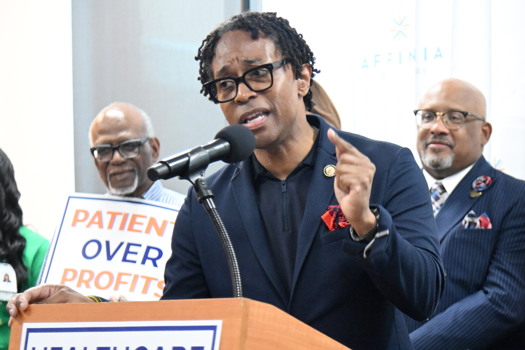 U.S. Rep. Wesley Bell speaks into a microphone at a podium next to a "Patients Over Profits" sign.