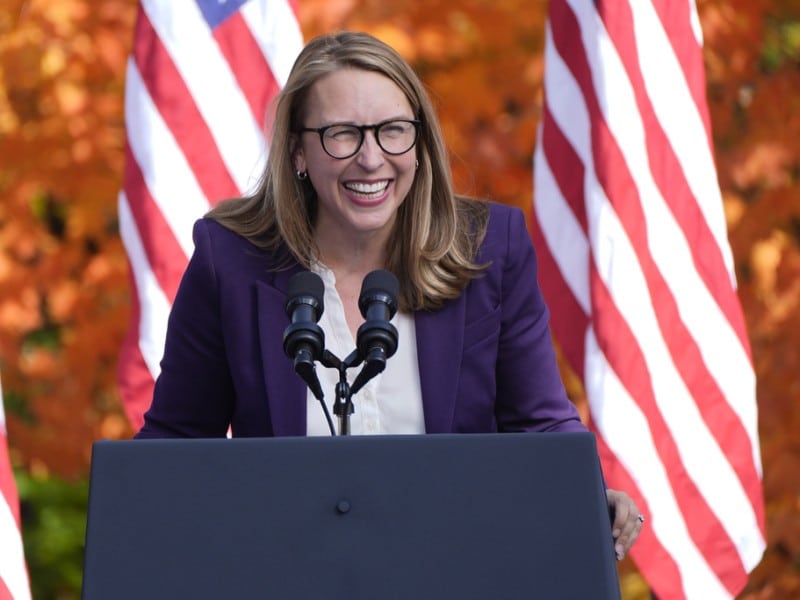 Rep. Hillary Scholten laughs while speaking at a podium in front of American flags and autumn foliage.