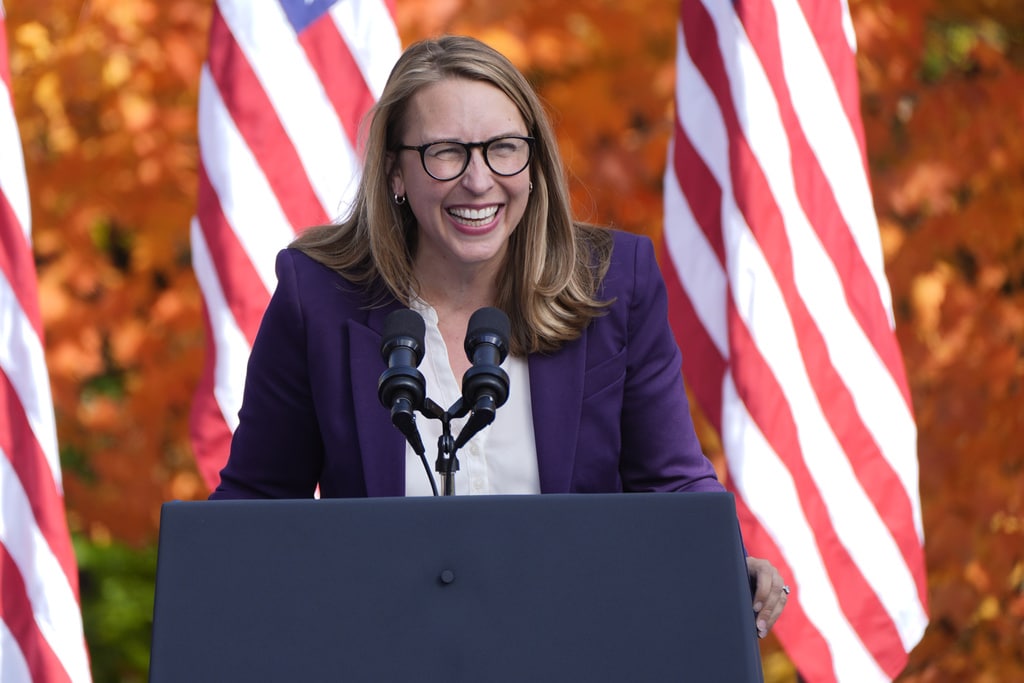 Rep. Hillary Scholten laughs while speaking at a podium in front of American flags and autumn foliage.