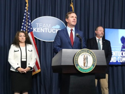 Gov. Andy Beshear speaks at a podium flanked by Angie Hatton and John Lyons in front of a "Team Kentucky" sign.