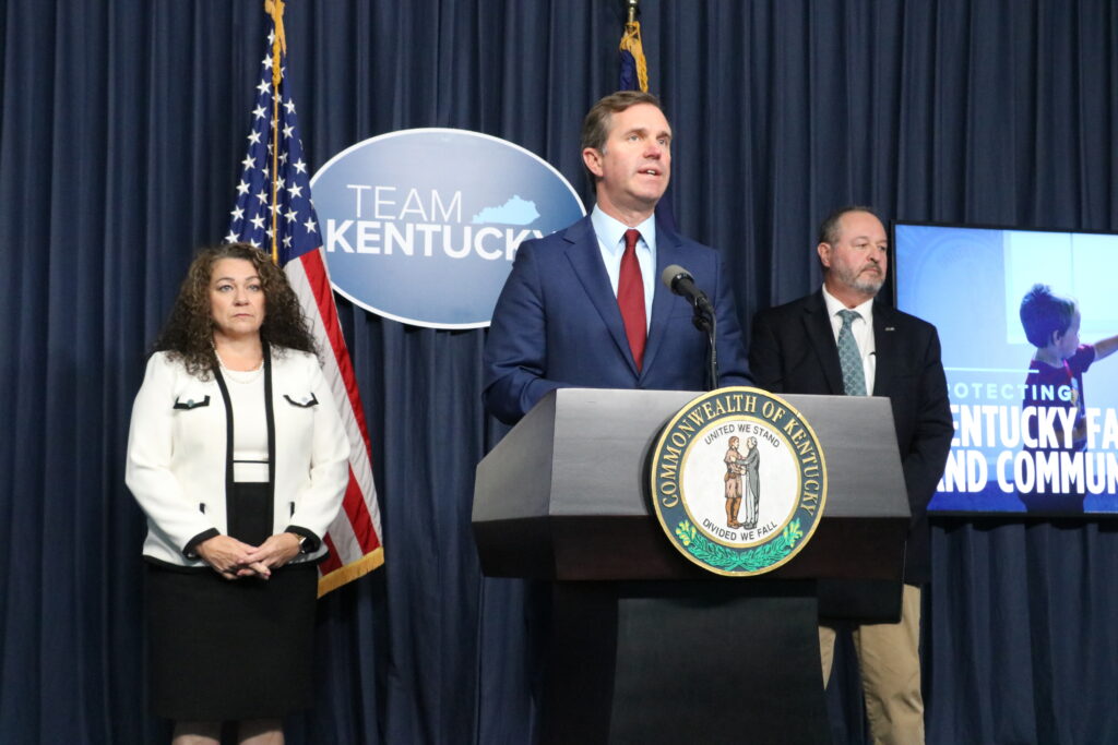 Gov. Andy Beshear speaks at a podium flanked by Angie Hatton and John Lyons in front of a "Team Kentucky" sign.