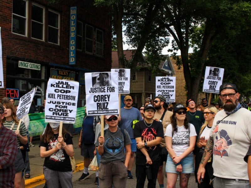 A crowd of demonstrators gathers outdoors holding signs reading "Justice for Corey Ruiz" and "Jail Killer Cop Kiel Peterson."