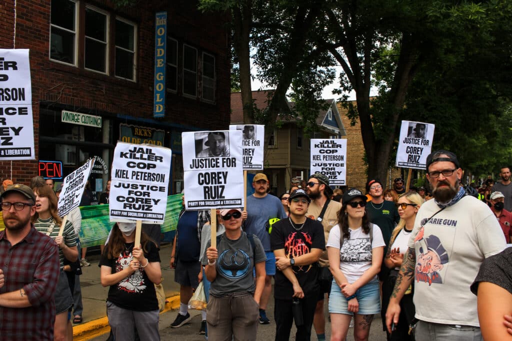 A crowd of demonstrators gathers outdoors holding signs reading "Justice for Corey Ruiz" and "Jail Killer Cop Kiel Peterson."