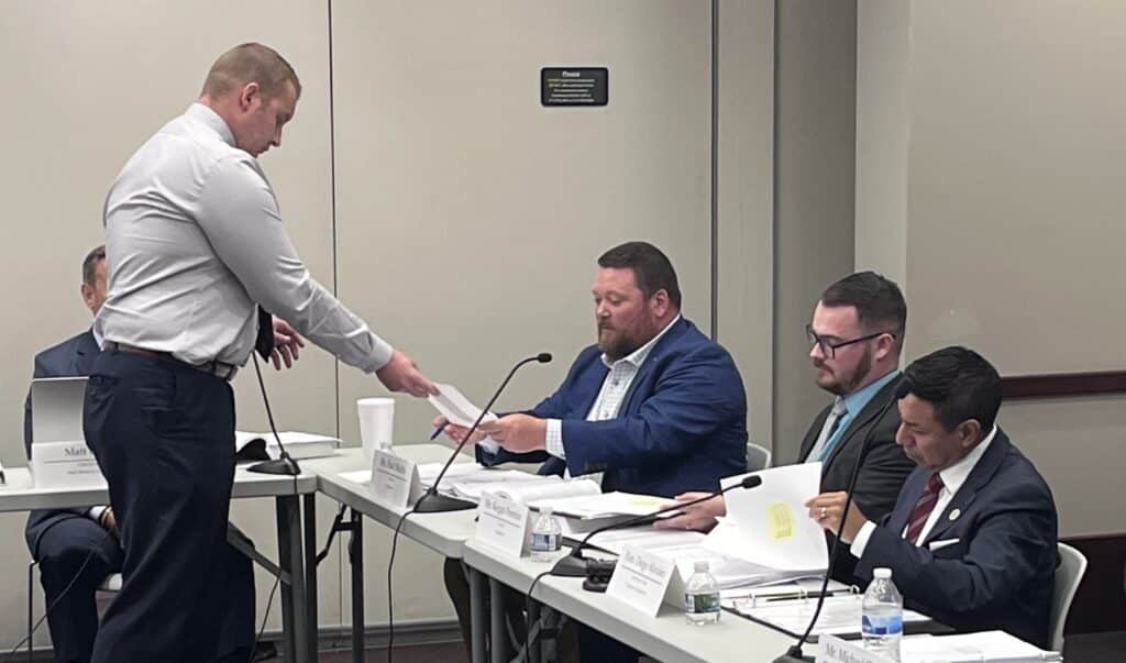 Evan Norris hands a paper to Paul Mullin, who sits at a panel table alongside other officials.