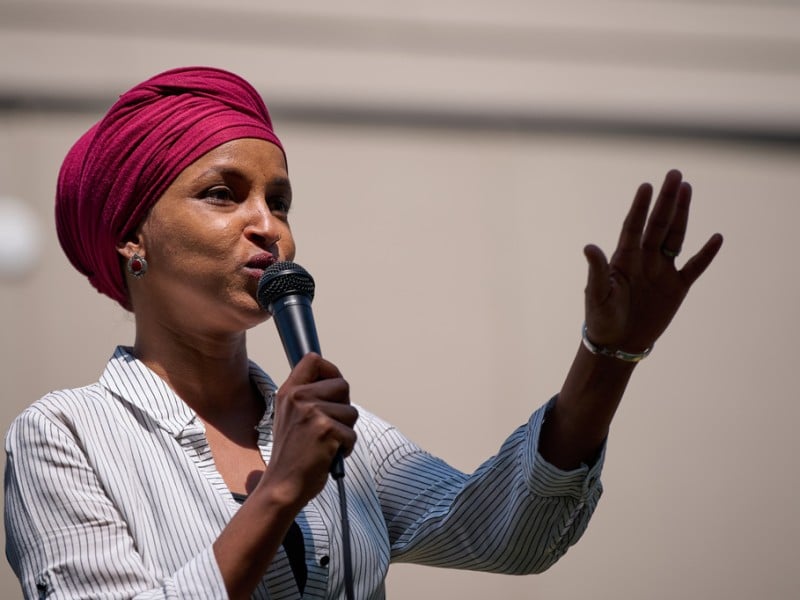 Rep. Ilhan Omar speaks into a microphone, gesturing with one hand raised while wearing a headscarf.