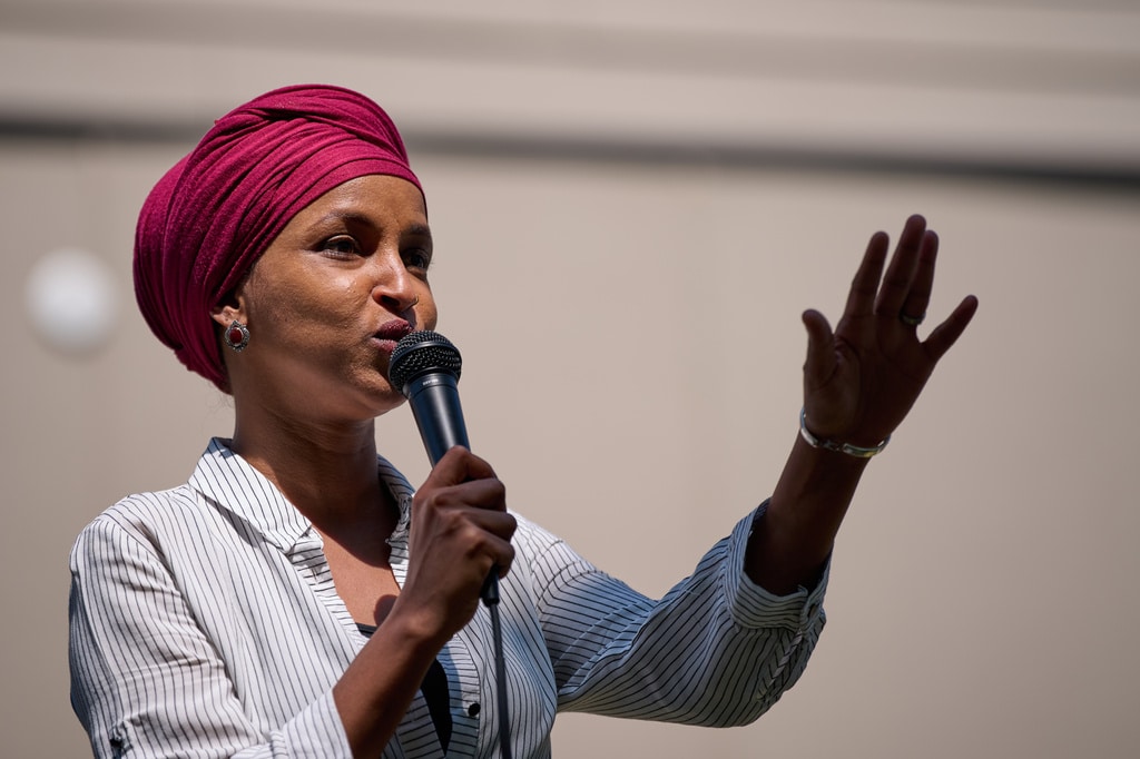 Rep. Ilhan Omar speaks into a microphone, gesturing with one hand raised while wearing a headscarf.