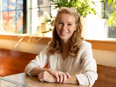 Rebecca Cooke smiles, sitting at a wooden table with her hands crossed in a brightly lit indoor space.