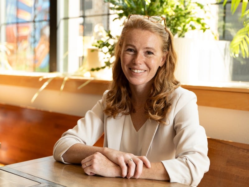 Rebecca Cooke smiles, sitting at a wooden table with her hands crossed in a brightly lit indoor space.