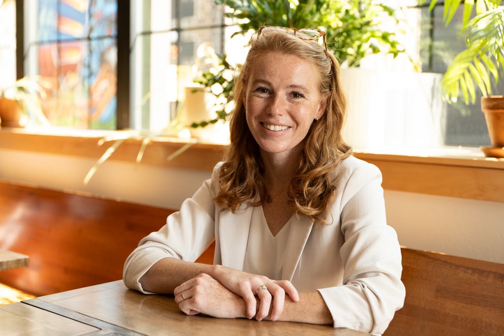 Rebecca Cooke smiles, sitting at a wooden table with her hands crossed in a brightly lit indoor space.