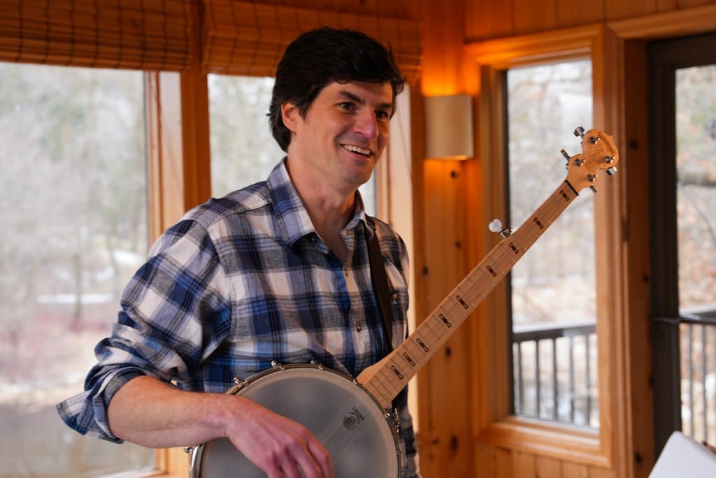 Matt Little smiles while holding a banjo indoors near a window.