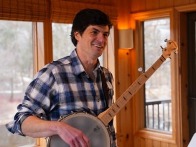 Matt Little smiles while holding a banjo indoors near a window.