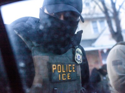 A masked officer wearing a vest labeled "POLICE ICE" looks through a car window.