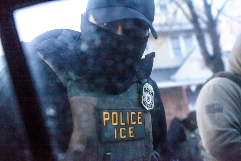 A masked officer wearing a vest labeled "POLICE ICE" looks through a car window.