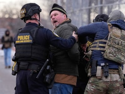 Homeland Security and tactical police officers restrain a shouting man outdoors near a fence.