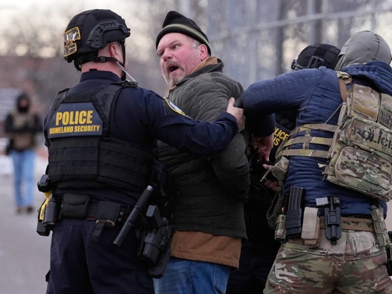 Homeland Security and tactical police officers restrain a shouting man outdoors near a fence.