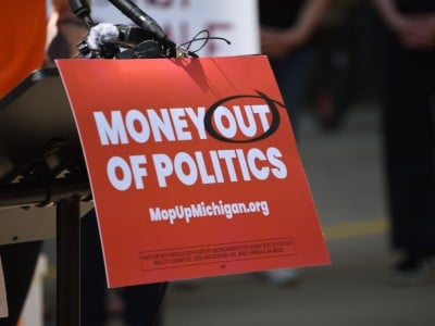 A podium sign reads "MONEY OUT OF POLITICS" with "OUT" circled, and lists "MopUpMichigan.org" below.
