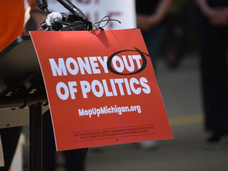 A podium sign reads "MONEY OUT OF POLITICS" with "OUT" circled, and lists "MopUpMichigan.org" below.