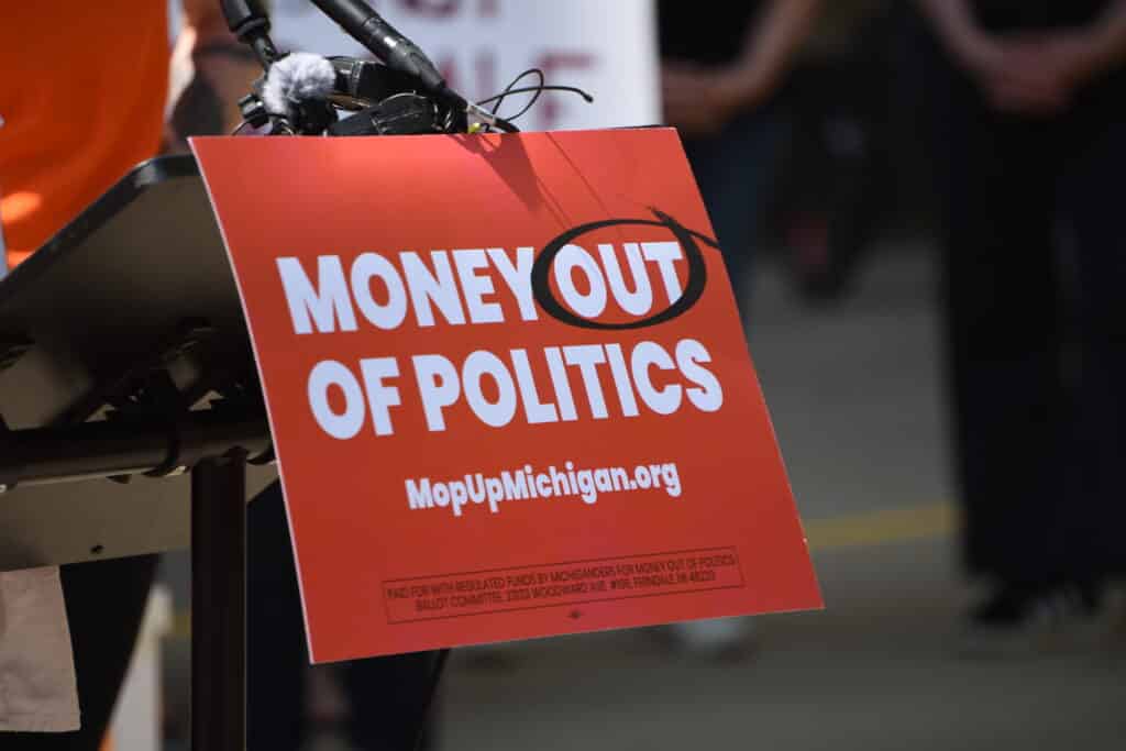 A podium sign reads "MONEY OUT OF POLITICS" with "OUT" circled, and lists "MopUpMichigan.org" below.