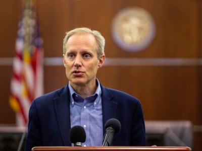 Steve Simon speaks at a wooden podium into microphones in front of an American flag and state seal.