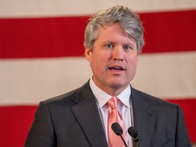 Nebraska Attorney General Mike Hilgers speaks into microphones in front of an American flag backdrop.