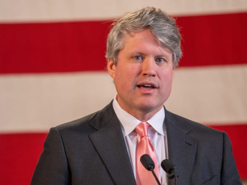 Nebraska Attorney General Mike Hilgers speaks into microphones in front of an American flag backdrop.