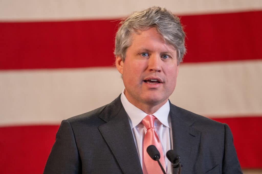 Nebraska Attorney General Mike Hilgers speaks into microphones in front of an American flag backdrop.