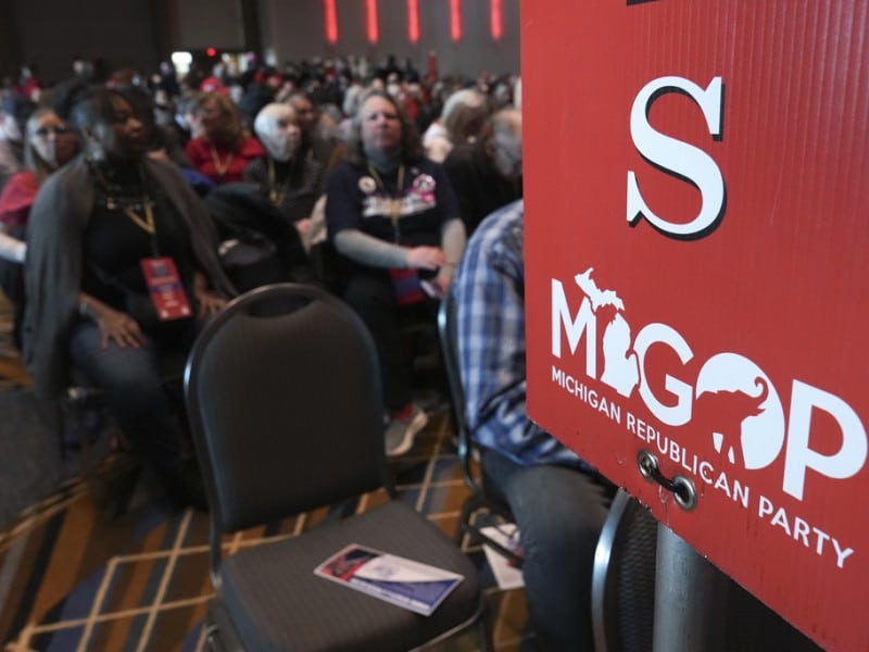 Delegates sit in a large hall behind a red sign displaying the letter "S" and the MIGOP Michigan Republican Party logo.