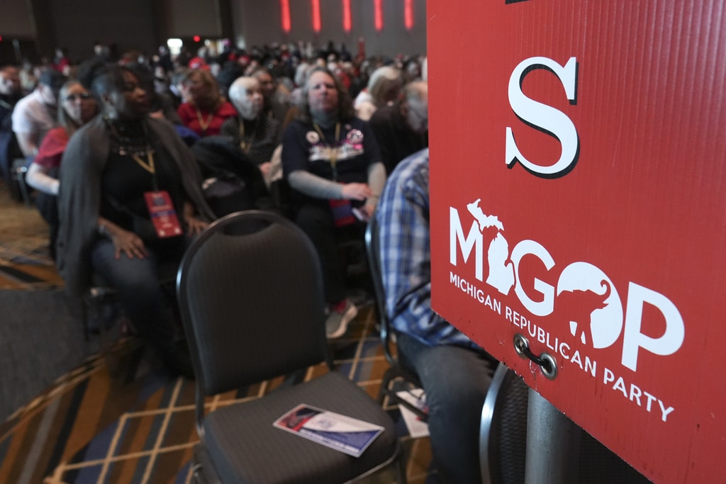 Delegates sit in a large hall behind a red sign displaying the letter "S" and the MIGOP Michigan Republican Party logo.