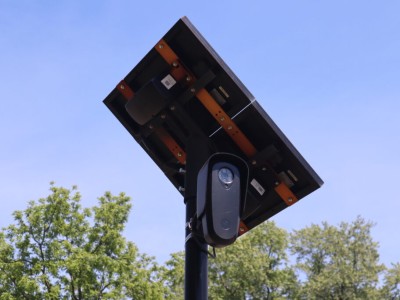 A Flock security camera with a small solar panel mounted above it on a pole against a clear sky.