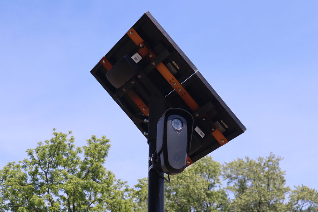 A Flock security camera with a small solar panel mounted above it on a pole against a clear sky.
