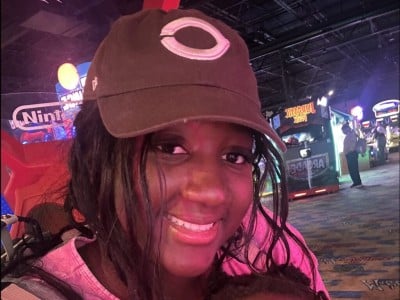 Armelle Bondonga smiles at the camera, wearing a baseball cap in an arcade with games lit up behind her.