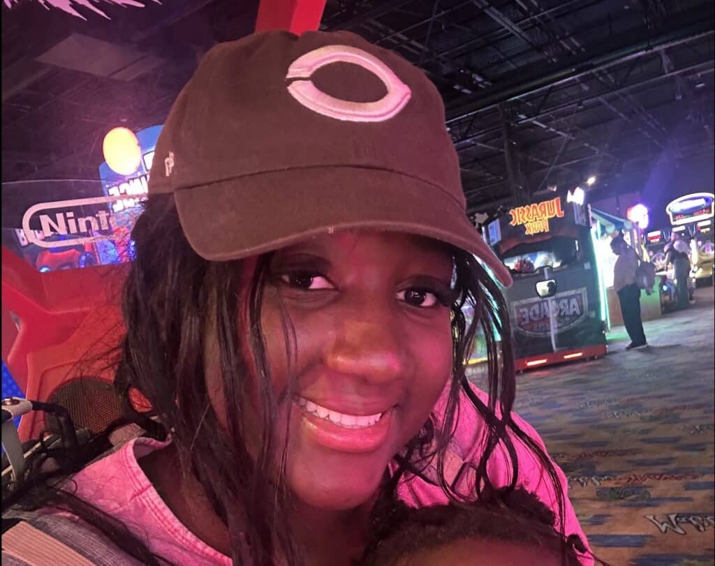 Armelle Bondonga smiles at the camera, wearing a baseball cap in an arcade with games lit up behind her.