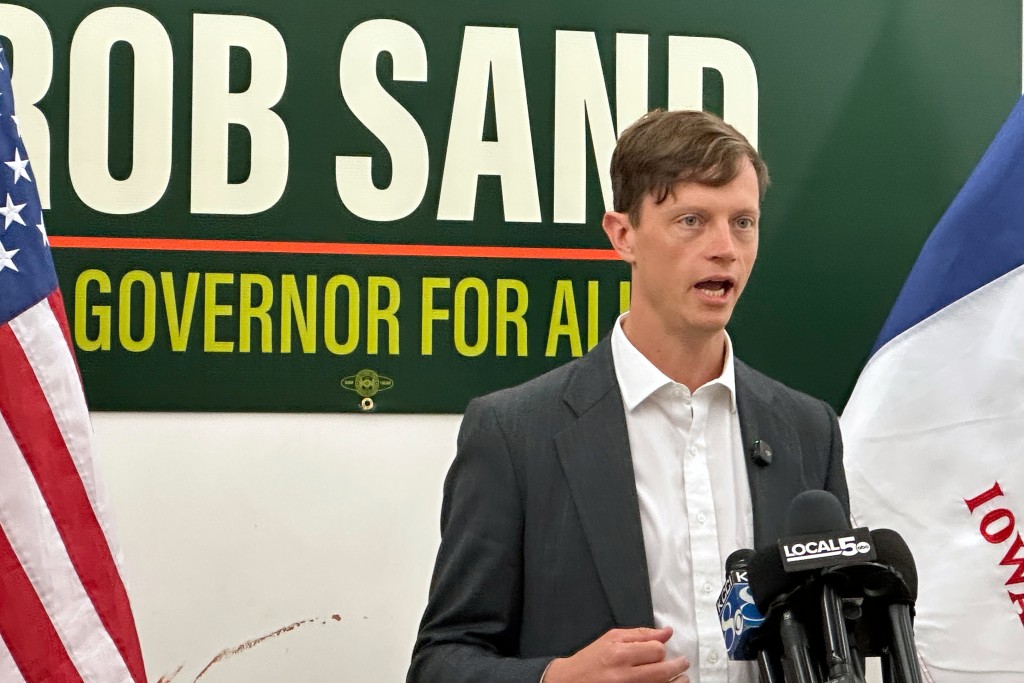 Rob Sand speaks into press microphones in front of a campaign sign reading "ROB SAND GOVERNOR FOR ALL."
