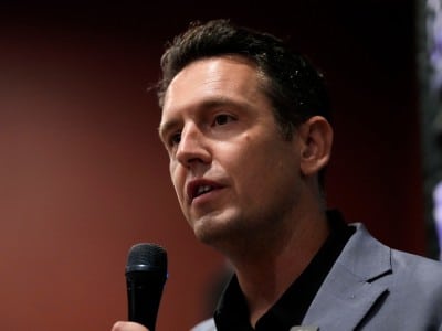 Zach Lahn speaks into a handheld microphone in a close-up profile view.