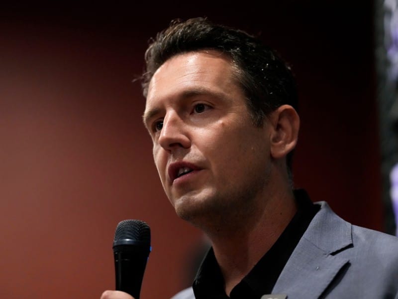 Zach Lahn speaks into a handheld microphone in a close-up profile view.
