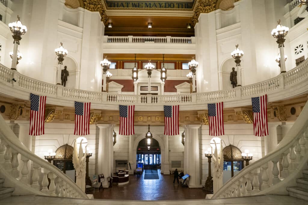 Inside the Capitol building: an ornate marble atrium with grand staircases, hanging American flags and a domed ceiling with gold accents.