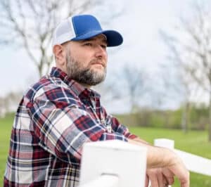 Brandon Dukes wearing a plaid shirt and cap rests his arms on a white fence in a grassy field.