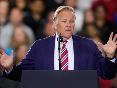 Mike Rogers speaks with open gestures at a podium in front of a crowded audience.