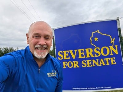 Erik Severson takes a selfie smiling next to a blue "SEVERSON FOR SENATE" campaign sign.