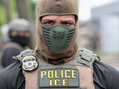 A close-up of a U.S. Immigration and Customs Enforcement officer wearing a balaclava, green facemask, and "POLICE ICE" vest.