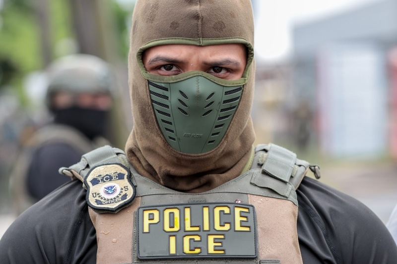 A close-up of a U.S. Immigration and Customs Enforcement officer wearing a balaclava, green facemask, and "POLICE ICE" vest.