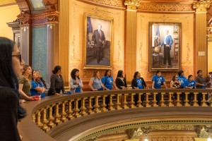 A group of people look down from an upper-level balcony inside an ornate, gilded Capitol building rotunda.