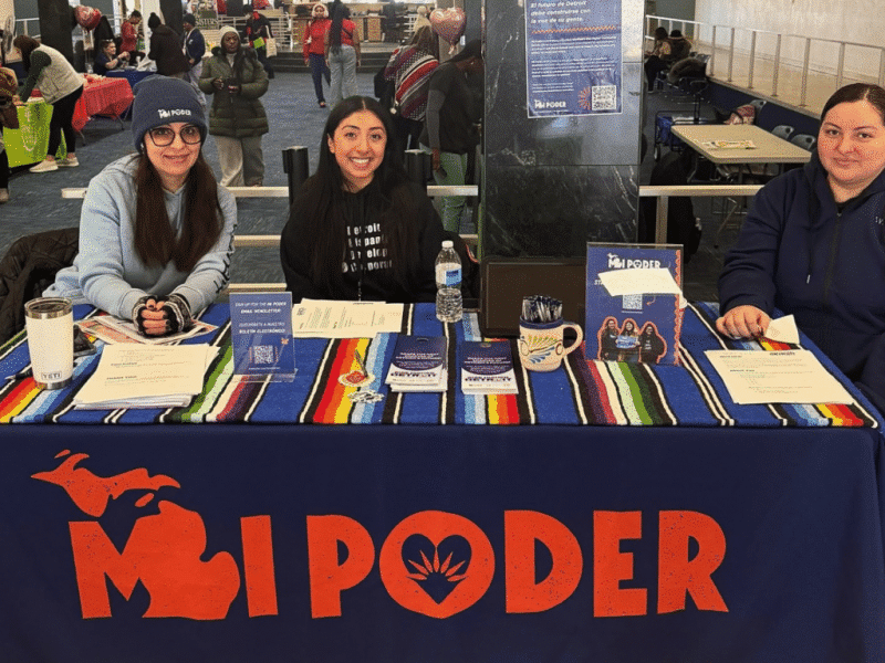 Three women sit behind a voter outreach table draped with a blue banner displaying a red "MI PODER" logo.