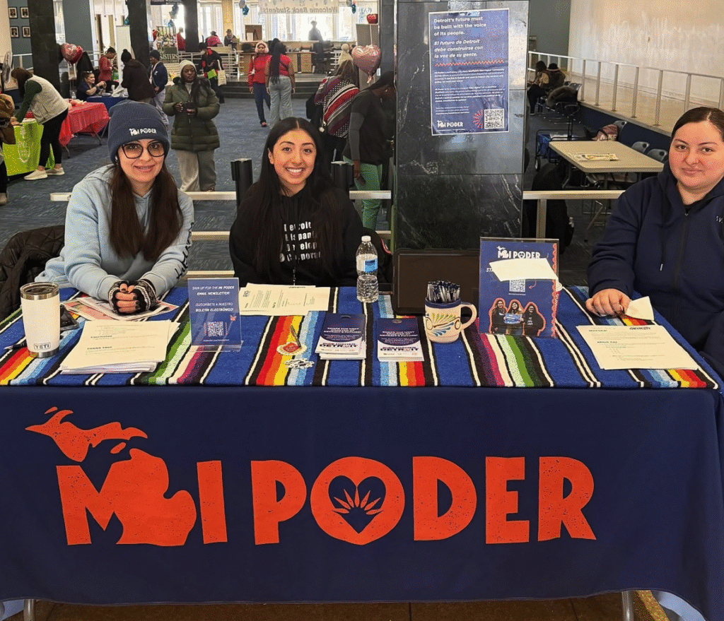 Three women sit behind a voter outreach table draped with a blue banner displaying a red "MI PODER" logo.