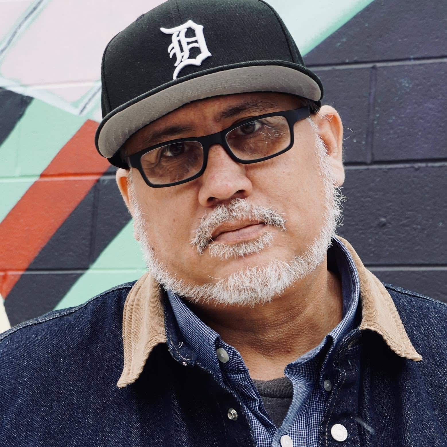 Pete Vargas wearing a Detroit Tigers cap, glasses and a denim jacket stands in front of a colorful wall.