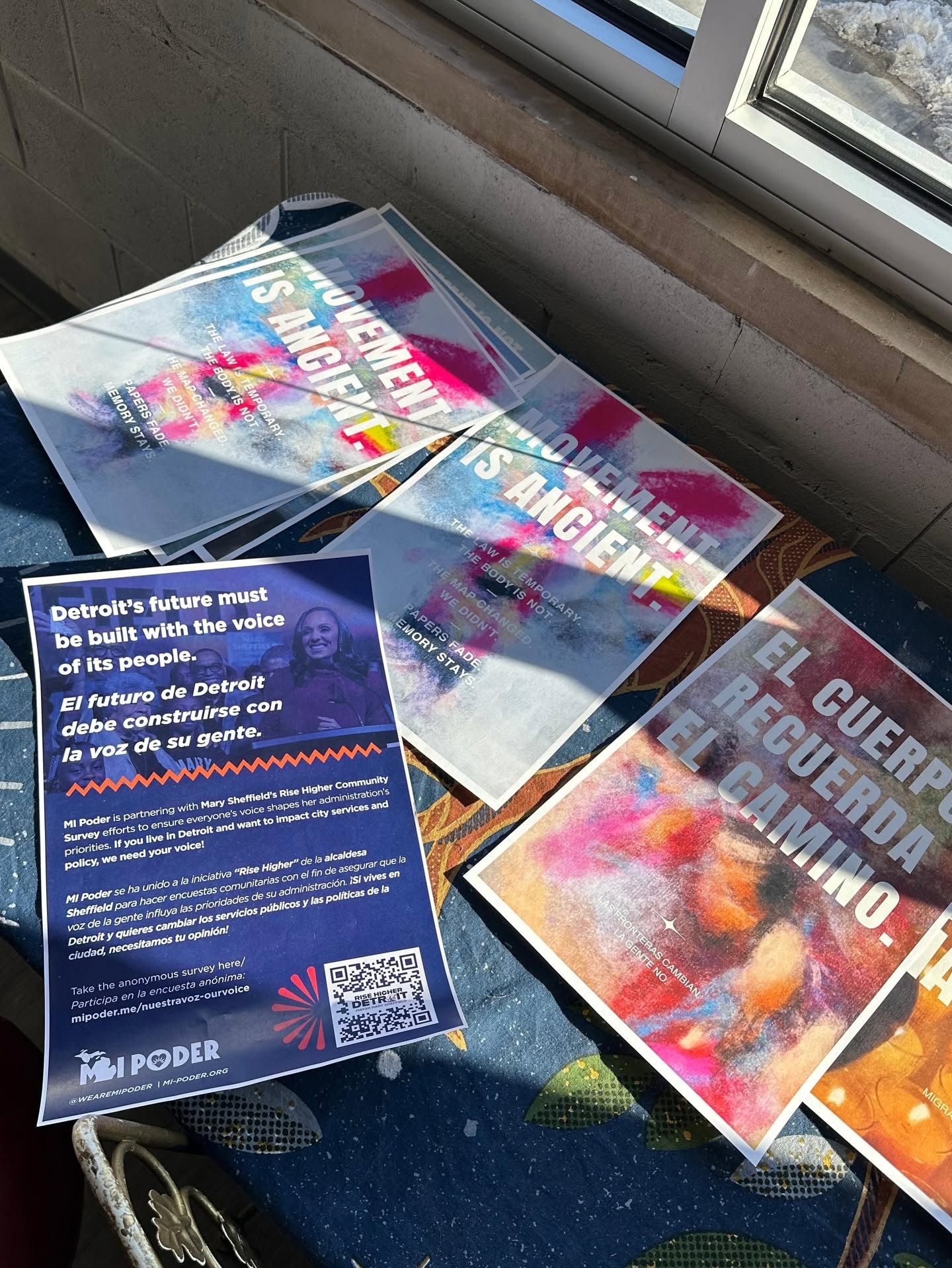 Flyers on a table read "MOVEMENT IS ANCIENT" and "EL CUERPO RECUERDA EL CAMINO" alongside an MI Poder survey handout.