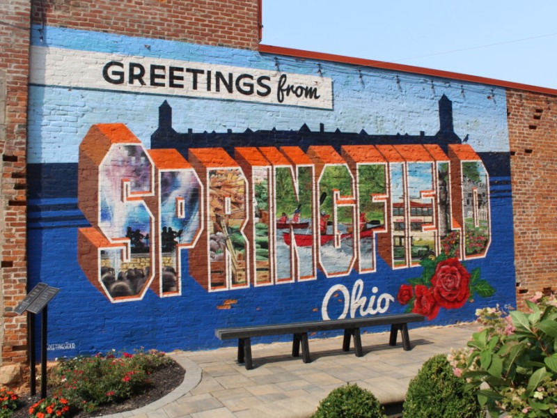 A brick building wall painted with a large mural reading "GREETINGS from SPRINGFIELD Ohio" in postcard style.